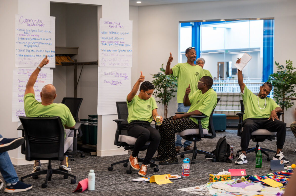 Restorative justice partnerships: Group of around 7 people all inlime green t-shirts sit rolling office chairs in a room with several poster paper tacked to walls filled with hand-written lists