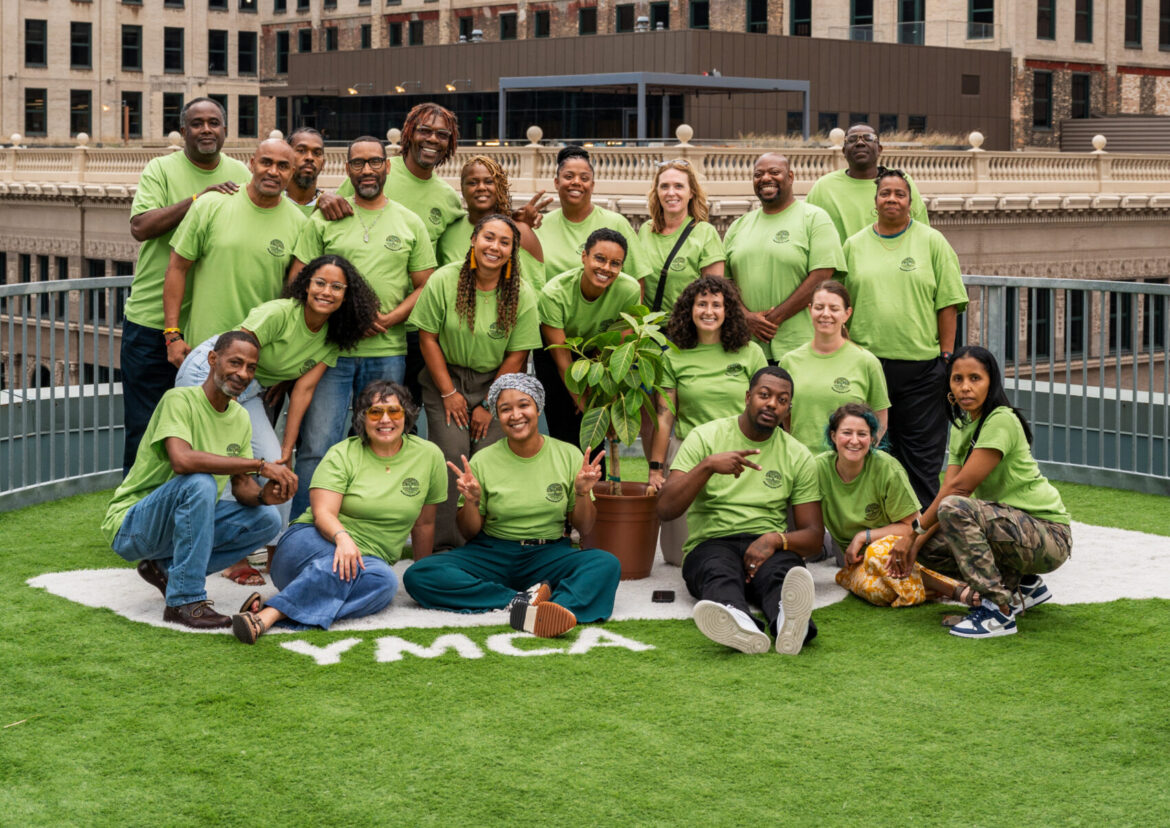 Restorative justice partnerships: Group of around 20 smiling people all wearing lime green t-shirts stand and sit in 3-4 rows on an artificial green turf field surrounded by tall city buildings