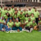 Restorative justice partnerships: Group of around 20 smiling people all wearing lime green t-shirts stand and sit in 3-4 rows on an artificial green turf field surrounded by tall city buildings