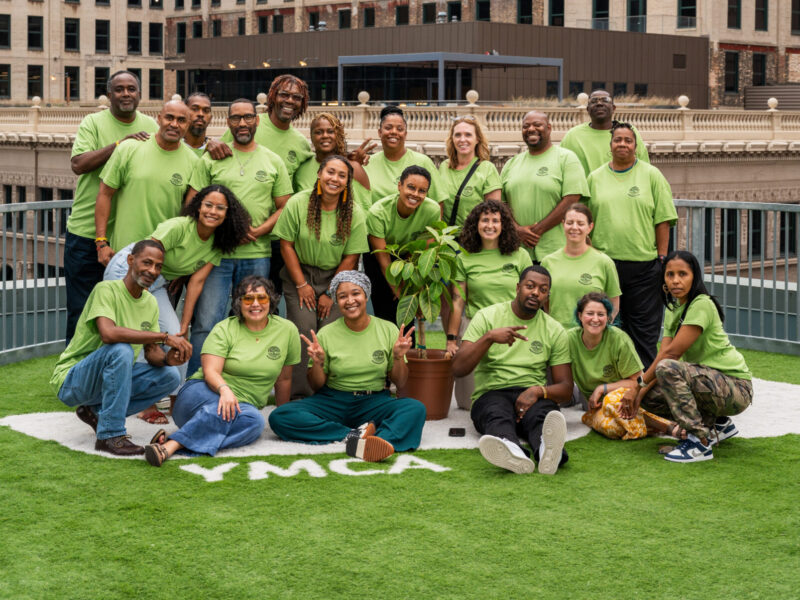 Restorative justice partnerships: Group of around 20 smiling people all wearing lime green t-shirts stand and sit in 3-4 rows on an artificial green turf field surrounded by tall city buildings