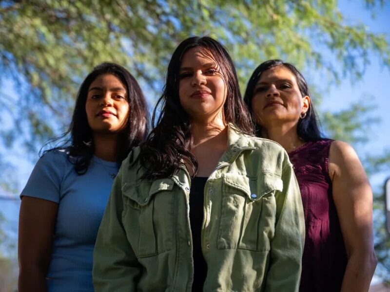 Latino gun reform Arizona: Thre Latino women with long, dark hair stand together facing camera with green tree foliage in the background